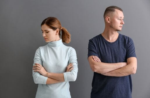 A man and woman, exhibiting the body language of discontented married couples, stand in front of a gray wall.