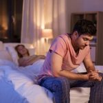 An image of a broken man sitting on a bed with his wife, both looking somber and lost in thought